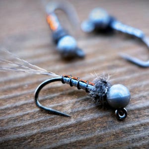May include: Close-up of three fishing flies. The foreground fly has a silver hook, a gray body, and a silver bead head. The body is wrapped with orange thread. The other two flies are blurred in the background.