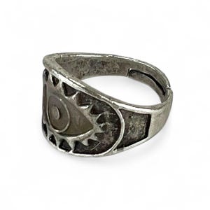 Puede incluir: Un anillo plateado con un diseño en forma de ojo. El anillo presenta una espiral central y formas triangulares circundantes. El anillo es ajustable. El anillo tiene un aspecto texturizado y antiguo.