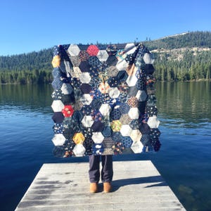 May include: A person is holding a large quilt made of hexagonal fabric patches in shades of blue, white, and red. The quilt is patterned with various designs and textures. The person is standing on a wooden jetty overlooking a lake with a forested shoreline.
