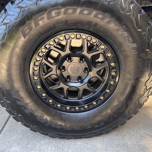 May include: Close-up of a black off-road vehicle wheel and tire. The tire is a BFGoodrich All-Terrain T/A KO2. The wheel is black with gold-colored accents. The wheel has a multi-spoke design.