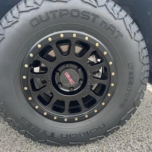 May include: A black automotive wheel with a matte finish, featuring a central GMC logo in red and silver, surrounded by a ring of small metallic bolts. The tire is a Nokian Outpost NAT model with the size LT 295/70R18 clearly visible on the sidewall.