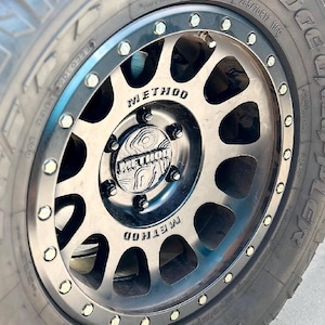 May include: Close-up of a black and silver Method wheel with a tire. The wheel has a series of oval cutouts and the word "METHOD" is printed on the wheel. The tire has the text "285/70R17 1160" printed on it.
