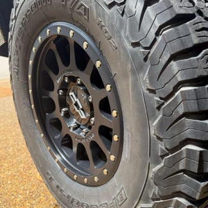 May include: A black off-road wheel rim with a multi-spoke design and a beadlock-style outer ring featuring silver bolts. The center cap displays the Method brand logo. The wheel is fitted with a BFGoodrich All-Terrain T/A KO2 tire with deep treads.