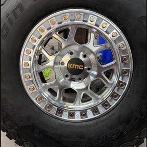 May include: Close-up of a black tire with a silver rim. The rim has a series of gold-colored bolts around the edge and a black center cap with the letters "KMC". The tire has the words "T/A KO2" printed on the side.