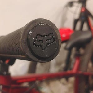 Puede incluir: Primer plano de un puño de manillar de bicicleta negro con una superficie texturizada. La tapa del extremo presenta el logotipo de Fox en negro. El fondo muestra un cuadro de bicicleta rojo y otras piezas de bicicleta.