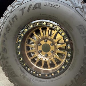 May include: A black BFGoodrich All-Terrain T/A KO2 tire mounted on a bronze-colored multi-spoke wheel. The wheel features a black outer beadlock ring secured with gold-toned bolts. The tire sidewall displays the text BFGoodrich and All-Terrain T/A KO2.
