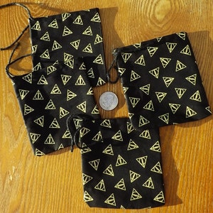 May include: Four black drawstring bags with a repeating gold triangle symbol pattern. The bags are arranged on a wooden surface. A dime is placed near the bags for size comparison. The bags are likely for small items.