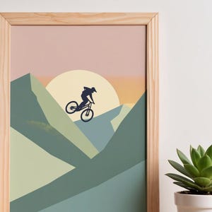 May include: A framed print featuring a mountain biker silhouetted against a sunset. The artwork uses a palette of muted greens, blues, and pinks, with a large yellow sun. The frame is made of light-colored wood. A succulent plant in a white pot is in the foreground.