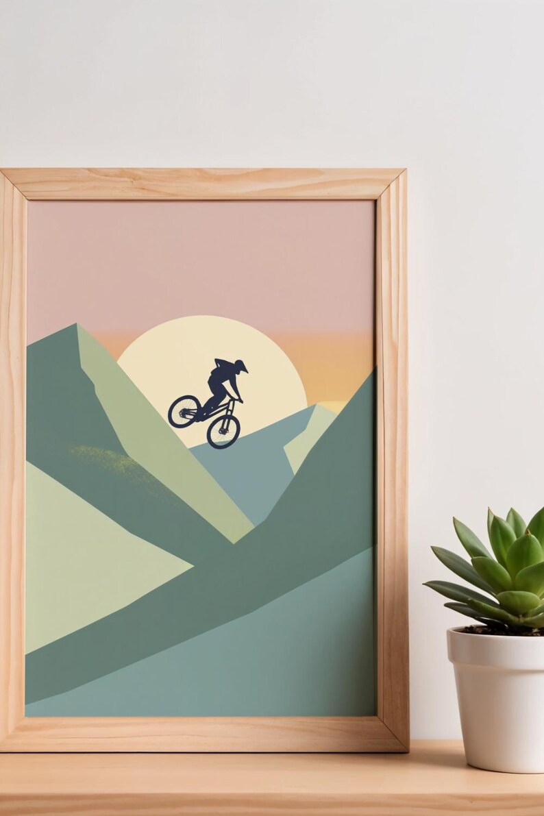 May include: A framed print featuring a mountain biker silhouetted against a sunset. The artwork uses a palette of muted greens, blues, and pinks, with a large yellow sun. The frame is made of light-colored wood. A succulent plant in a white pot is in the foreground.