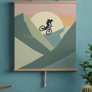 May include: A mountain biking art print featuring a silhouette of a cyclist mid-air against a sunset backdrop. The artwork uses shades of green, peach, and orange, and is displayed with wooden bars and a hanging string. The theme is adventure and outdoor sports.