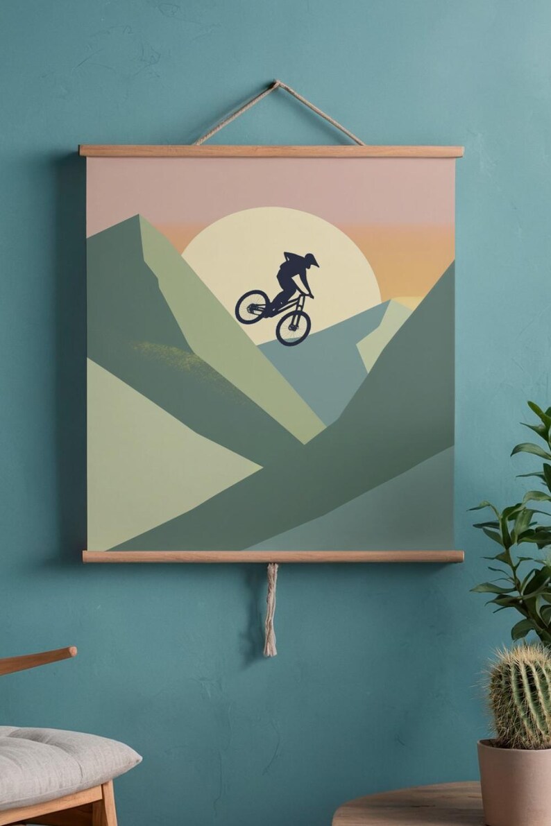 May include: A mountain biking art print featuring a silhouette of a cyclist mid-air against a sunset backdrop. The artwork uses shades of green, peach, and orange, and is displayed with wooden bars and a hanging string. The theme is adventure and outdoor sports.