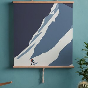 May include: A mountain landscape art print featuring a skier ascending a snow-covered mountain. The artwork is in shades of blue and white, with a dark blue sky. The print is held by wooden bars and a natural rope.