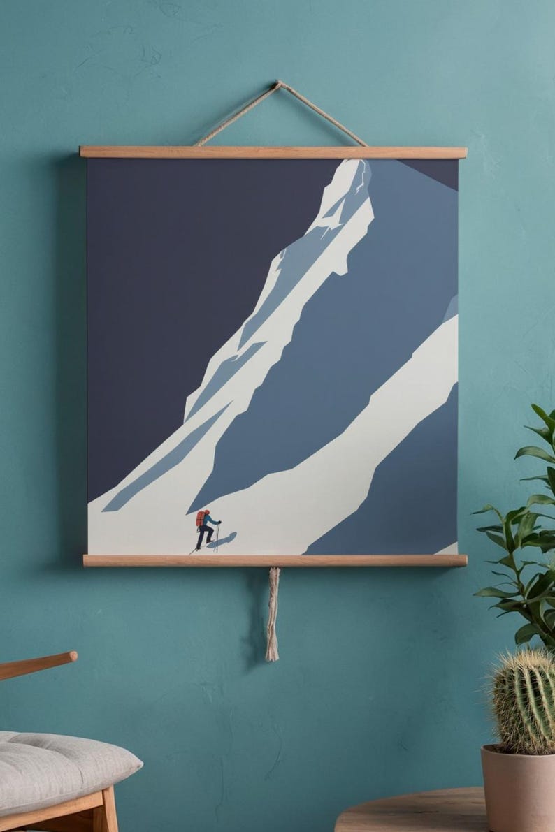 May include: A mountain landscape art print featuring a skier ascending a snow-covered mountain. The artwork is in shades of blue and white, with a dark blue sky. The print is held by wooden bars and a natural rope.