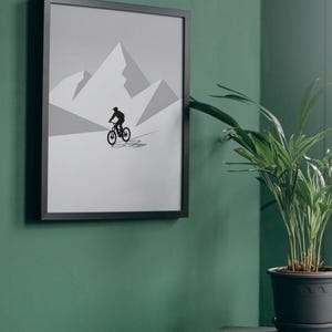 May include: Framed art print featuring a black silhouette of a mountain biker on a gray and white mountain landscape. The artwork is displayed on a green wall, with a potted plant in the foreground. The frame is black.