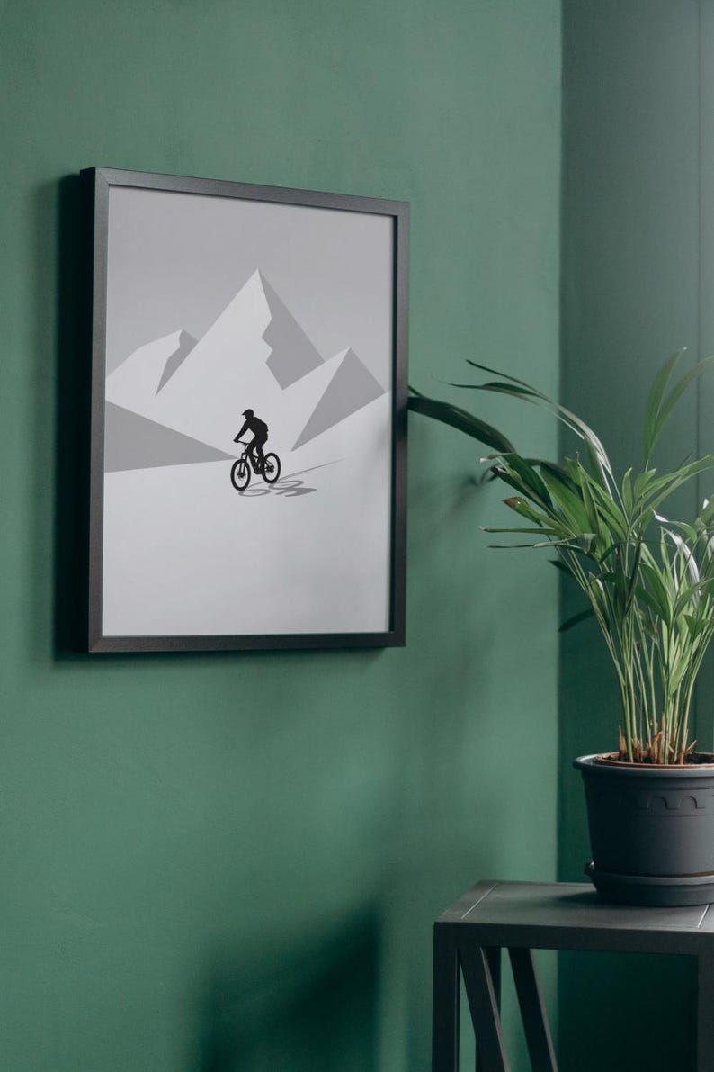 May include: Framed art print featuring a black silhouette of a mountain biker on a gray and white mountain landscape. The artwork is displayed on a green wall, with a potted plant in the foreground. The frame is black.