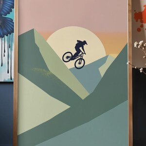 May include: A framed art print featuring a mountain biker silhouetted against a sunset. The artwork uses a palette of muted greens, pinks, and oranges to depict a mountain landscape. The print is encased in a wooden frame.