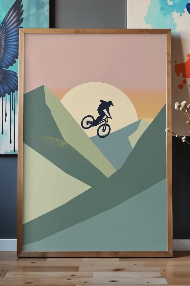 May include: A framed art print featuring a mountain biker silhouetted against a sunset. The artwork uses a palette of muted greens, pinks, and oranges to depict a mountain landscape. The print is encased in a wooden frame.