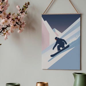 May include: A framed wall art print featuring a snowboarder in a black silhouette, descending a snowy mountain slope. The artwork uses a minimalist style with shades of blue, pink, and white. The print is suspended by a wooden dowel and a natural cord.