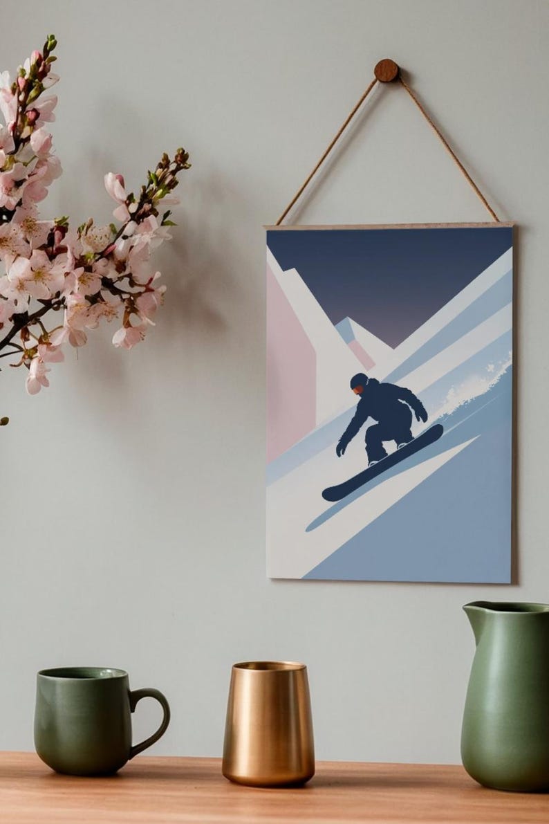 May include: A framed wall art print featuring a snowboarder in a black silhouette, descending a snowy mountain slope. The artwork uses a minimalist style with shades of blue, pink, and white. The print is suspended by a wooden dowel and a natural cord.