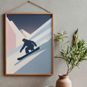 May include: A framed art print featuring a snowboarder carving down a snowy mountain slope. The artwork uses a minimalist style with shades of blue, pink, and white, and is displayed with a natural wood frame and a hanging cord.