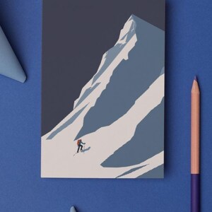 May include: A rectangular print depicts a skier descending a snow-covered mountain. The artwork features a minimalist style with a color palette of blues, white, and a touch of red for the skier's backpack. A pencil and paper cutouts are on a blue surface.