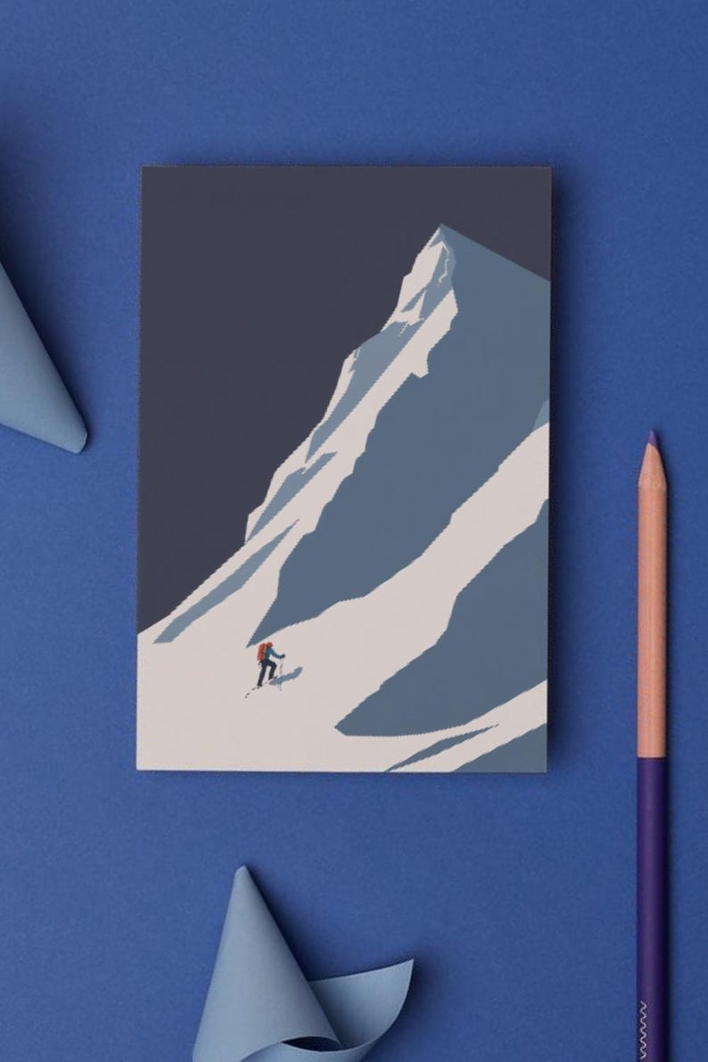 May include: A rectangular print depicts a skier descending a snow-covered mountain. The artwork features a minimalist style with a color palette of blues, white, and a touch of red for the skier's backpack. A pencil and paper cutouts are on a blue surface.