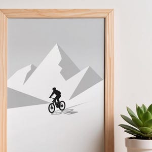 May include: Framed art print featuring a black silhouette of a mountain biker riding down a slope, set against a backdrop of stylized, geometric mountain peaks in shades of gray. The artwork is in a light wood frame.