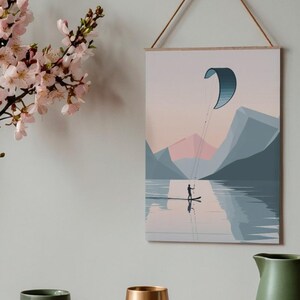 May include: A framed art print depicting a person kitesurfing on a lake, with mountains in the background. The artwork features a minimalist style with a color palette of blues, pinks, and grays. The print is hung by a wooden dowel and a natural cord.