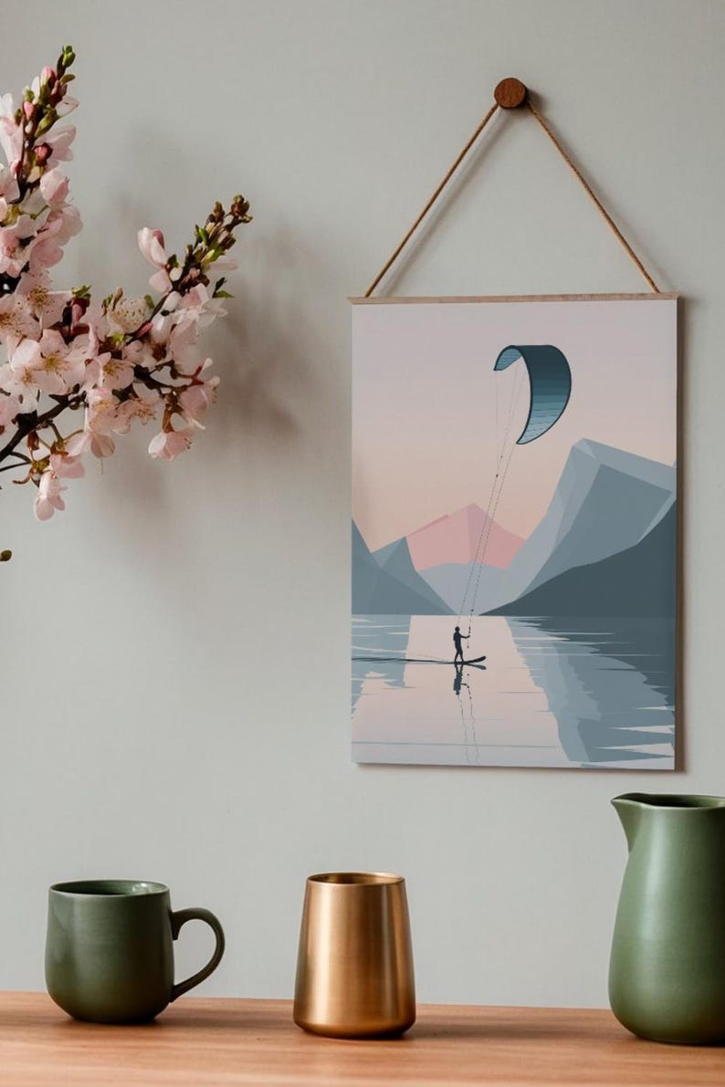 May include: A framed art print depicting a person kitesurfing on a lake, with mountains in the background. The artwork features a minimalist style with a color palette of blues, pinks, and grays. The print is hung by a wooden dowel and a natural cord.