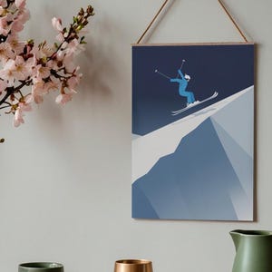 May include: A framed wall art print featuring a skier in a blue jumpsuit and white helmet, mid-air on a snowy mountain slope. The artwork is minimalist with a dark blue sky and white snow. The print is suspended by a wooden bar and a natural rope.