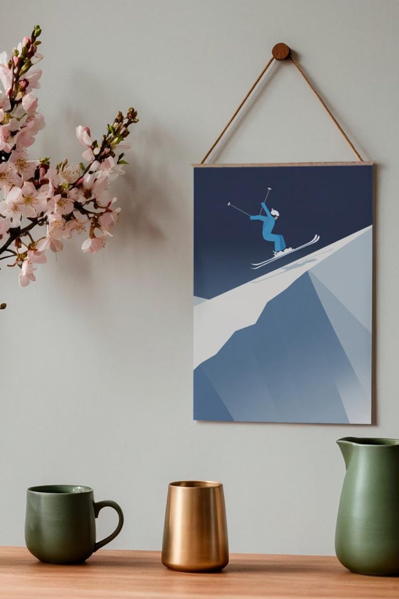May include: A framed wall art print featuring a skier in a blue jumpsuit and white helmet, mid-air on a snowy mountain slope. The artwork is minimalist with a dark blue sky and white snow. The print is suspended by a wooden bar and a natural rope.