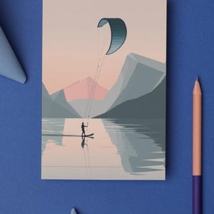 May include: A postcard featuring a watersports scene. The illustration depicts a person kitesurfing on a lake with mountains in the background. The color palette includes shades of blue, pink, and gray. A pencil and paper are on a blue surface.