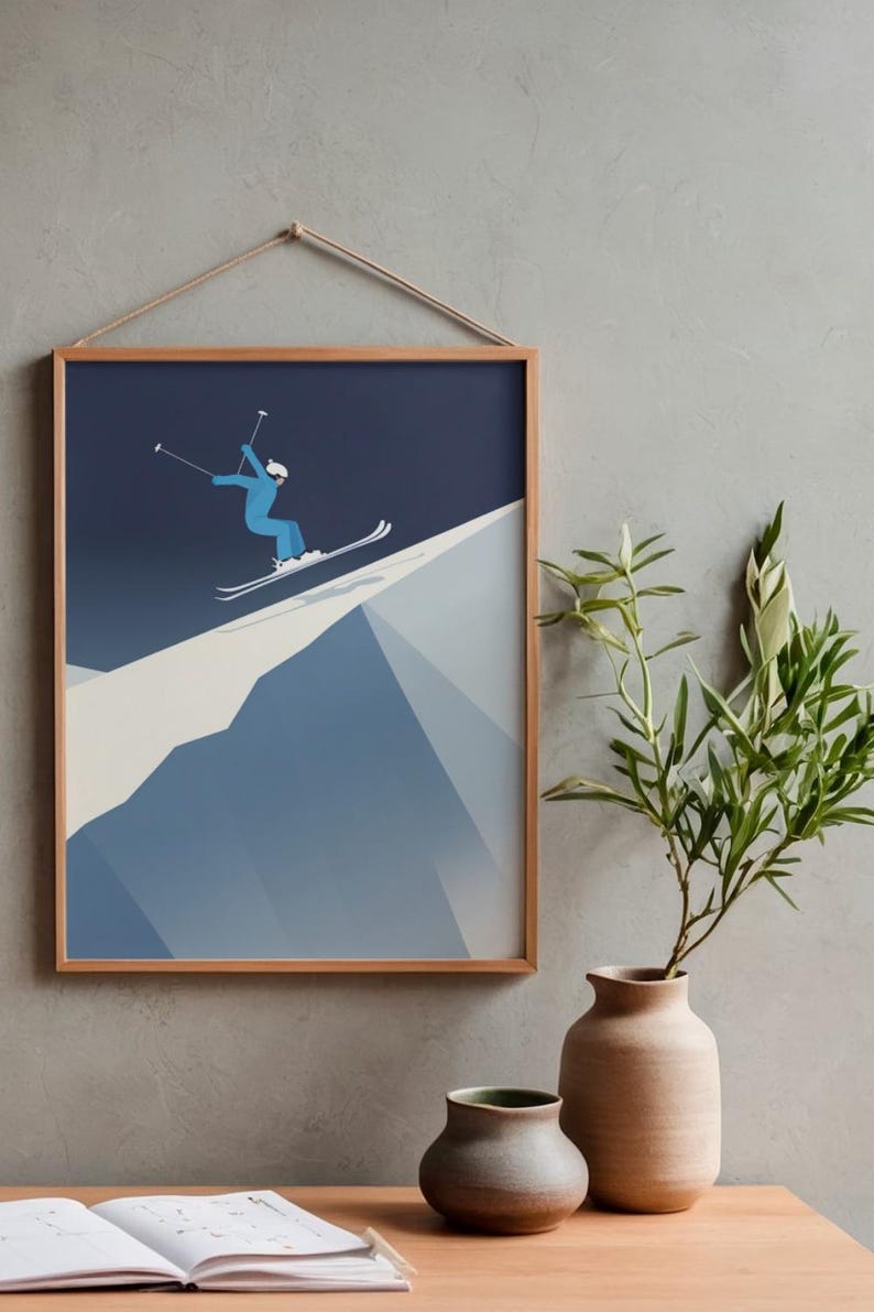 May include: A framed print depicts a skier in a blue suit and white helmet skiing down a mountain. The artwork is in a minimalist style with a dark blue sky and a light blue and white mountain. The frame is made of light brown wood.