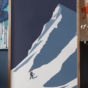 May include: Framed art print featuring a minimalist illustration of a mountain climber ascending a snow-covered peak. The artwork uses a palette of blues and white, with a dark blue sky. The frame is a light brown color.