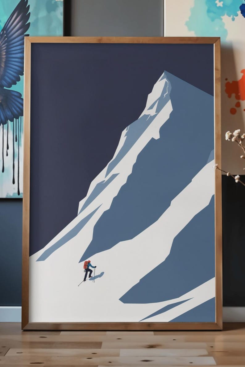 May include: Framed art print featuring a minimalist illustration of a mountain climber ascending a snow-covered peak. The artwork uses a palette of blues and white, with a dark blue sky. The frame is a light brown color.
