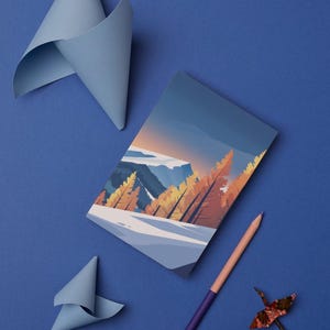 May include: A notebook with a landscape illustration, a purple and peach pencil, and origami paper cranes are arranged on a blue surface. Two folded paper shapes in light blue are also present. The notebook features a mountain scene.