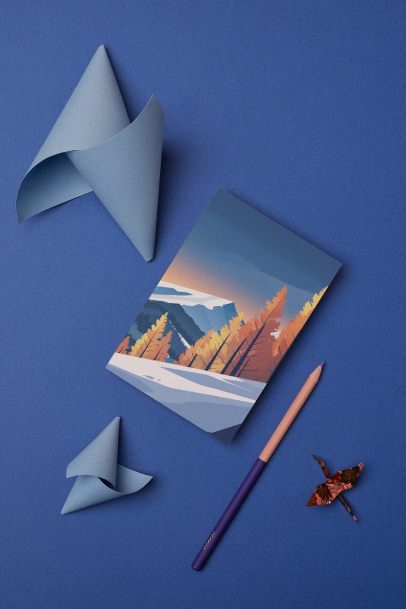 May include: A notebook with a landscape illustration, a purple and peach pencil, and origami paper cranes are arranged on a blue surface. Two folded paper shapes in light blue are also present. The notebook features a mountain scene.