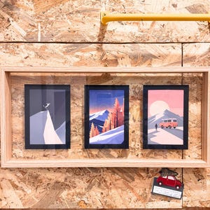 May include: Three framed art prints with mountain and travel themes. The prints feature a skier, a mountain landscape with trees, and a van on a road. The frames are light wood, and the background is a wood composite.