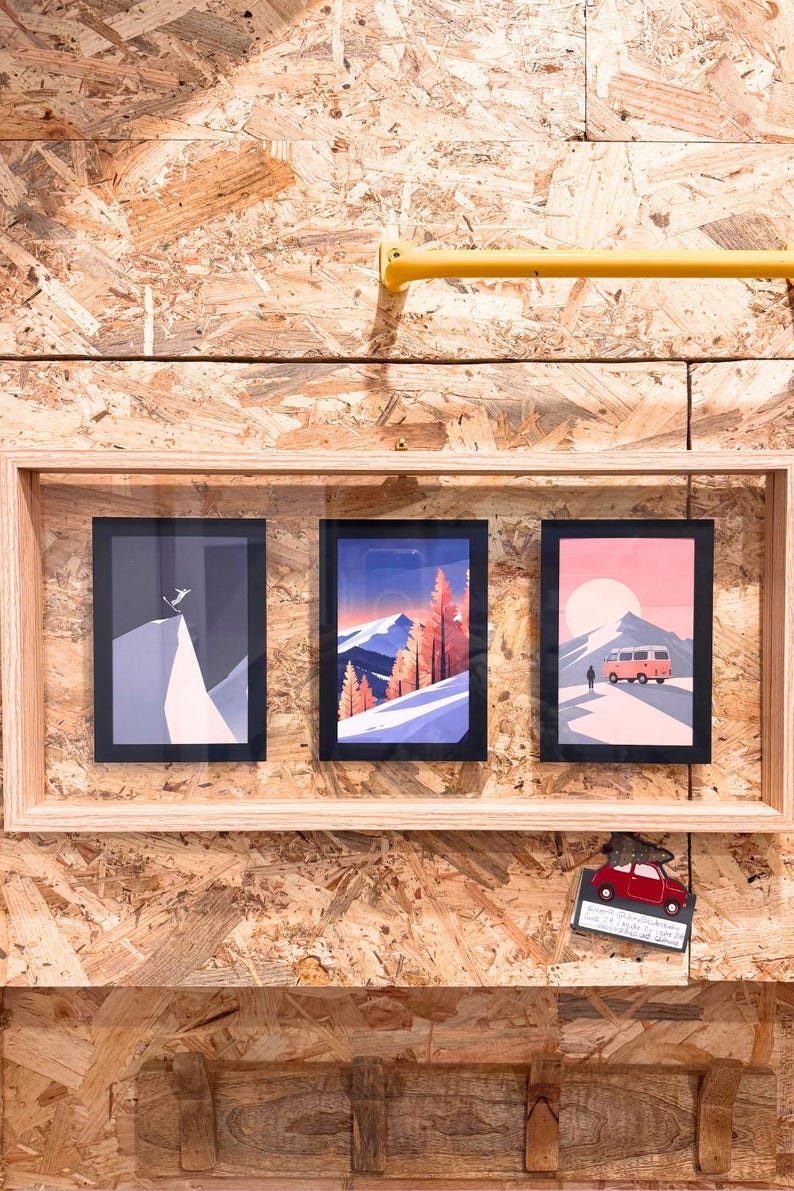 May include: Three framed art prints with mountain and travel themes. The prints feature a skier, a mountain landscape with trees, and a van on a road. The frames are light wood, and the background is a wood composite.