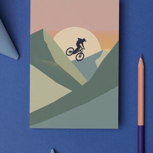 May include: A mountain biking-themed art print. The illustration features a cyclist silhouetted against a sunset over a mountain range. The color palette includes shades of blue, green, and pink. A purple and peach colored pencil is on the blue background.