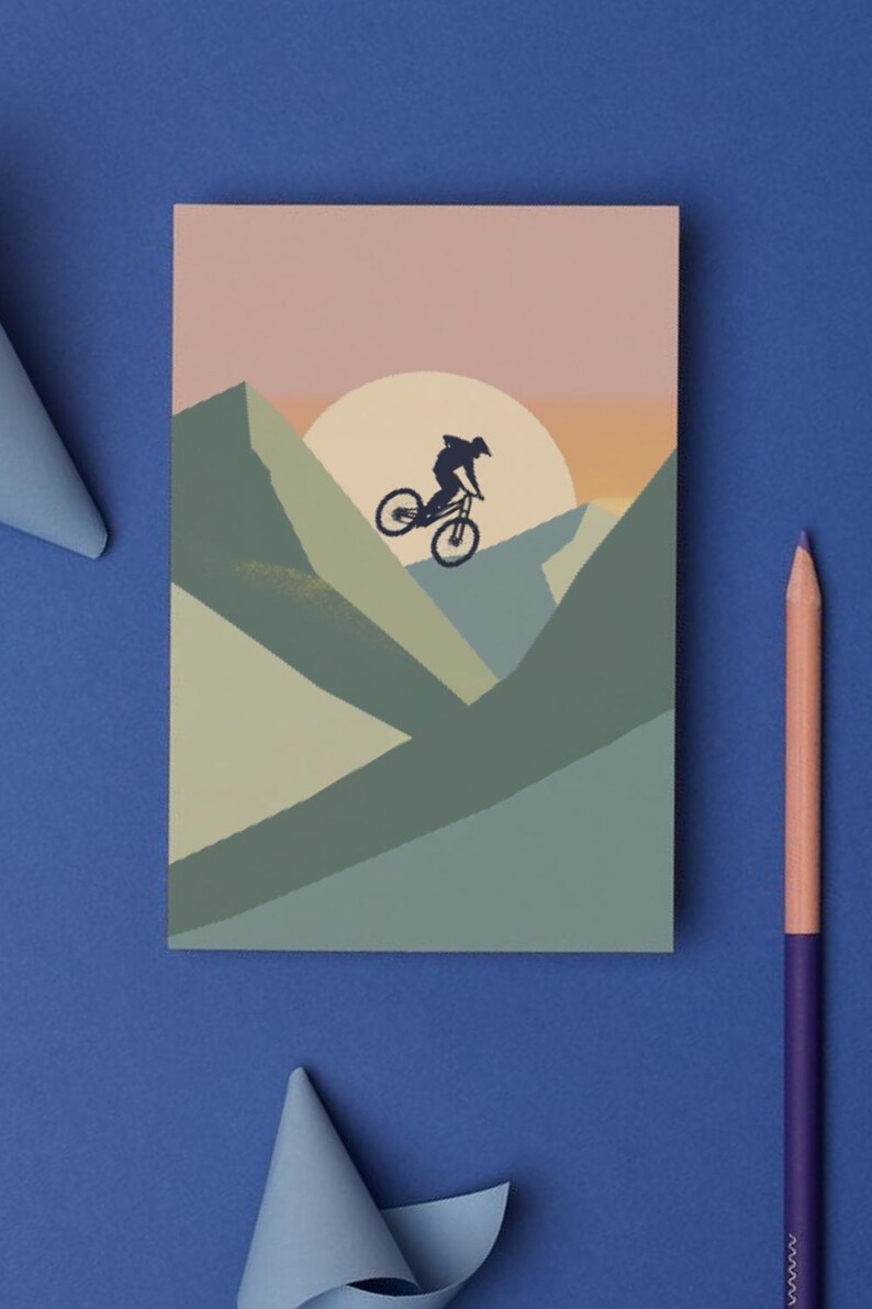 May include: A mountain biking-themed art print. The illustration features a cyclist silhouetted against a sunset over a mountain range. The color palette includes shades of blue, green, and pink. A purple and peach colored pencil is on the blue background.