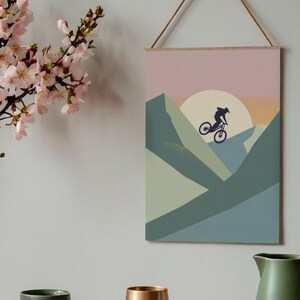 May include: A framed wall art print featuring a mountain biking scene. The artwork depicts a cyclist silhouetted against a sunset, with geometric mountain shapes in shades of green and blue. The print is suspended by a wooden dowel and a natural cord.