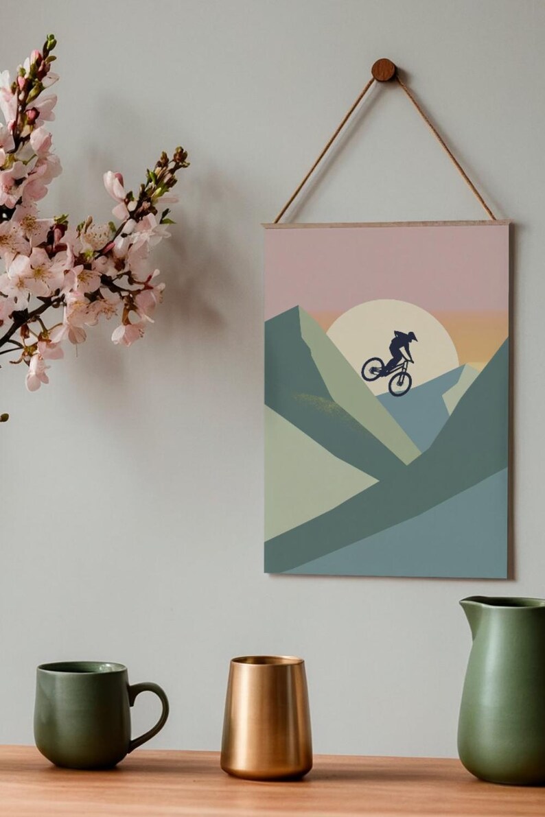 May include: A framed wall art print featuring a mountain biking scene. The artwork depicts a cyclist silhouetted against a sunset, with geometric mountain shapes in shades of green and blue. The print is suspended by a wooden dowel and a natural cord.