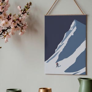 May include: A framed art print depicting a skier descending a snow-covered mountain. The artwork features a minimalist style with shades of blue and white. The print is suspended by a wooden bar and a natural cord. A green mug, a gold vase, and a green pitcher are in the foreground.
