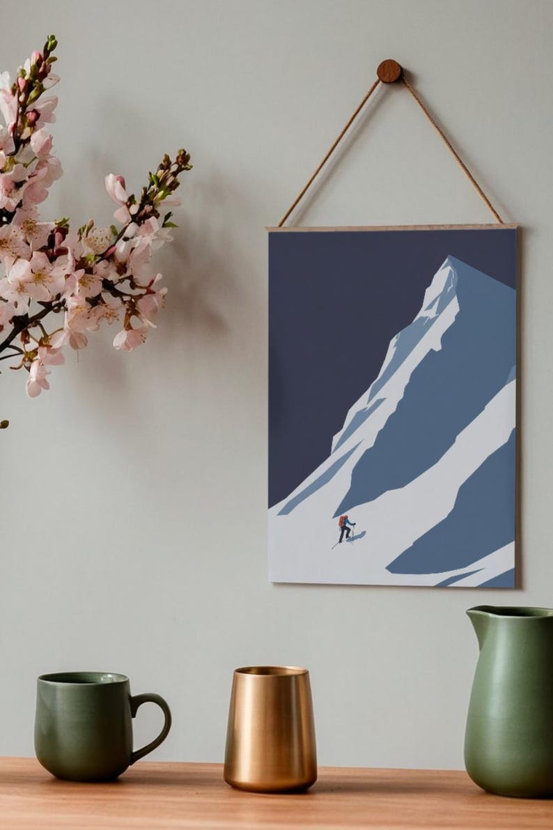 May include: A framed art print depicting a skier descending a snow-covered mountain. The artwork features a minimalist style with shades of blue and white. The print is suspended by a wooden bar and a natural cord. A green mug, a gold vase, and a green pitcher are in the foreground.