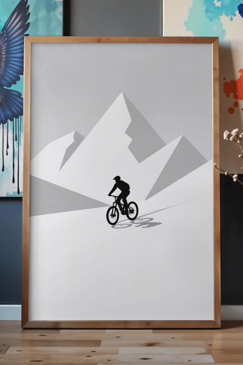May include: A framed print featuring a minimalist illustration of a mountain biker silhouetted against a backdrop of stylized mountain peaks. The artwork is primarily in shades of gray and white, with a wooden frame.