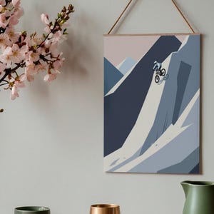 May include: A framed print depicts a mountain biker descending a steep, snow-covered mountain. The artwork uses shades of blue, gray, and white. The print is suspended by a wooden dowel and a natural fiber cord. A sprig of pink flowers is in the upper left.