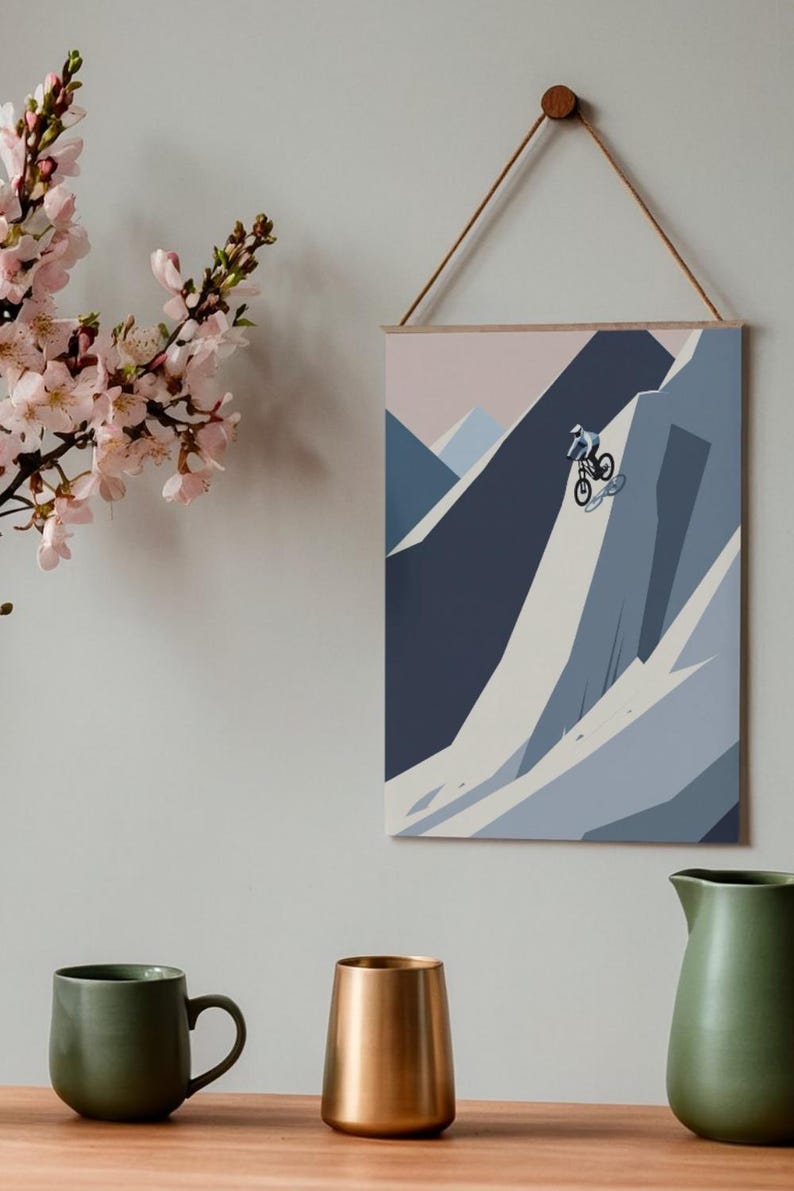 May include: A framed print depicts a mountain biker descending a steep, snow-covered mountain. The artwork uses shades of blue, gray, and white. The print is suspended by a wooden dowel and a natural fiber cord. A sprig of pink flowers is in the upper left.