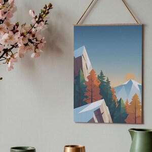 May include: A framed art print featuring a geometric mountain landscape with trees in shades of green, orange, and brown. The artwork is suspended by a wooden dowel and a natural cord. The background is a gradient of blue and the sun is setting.