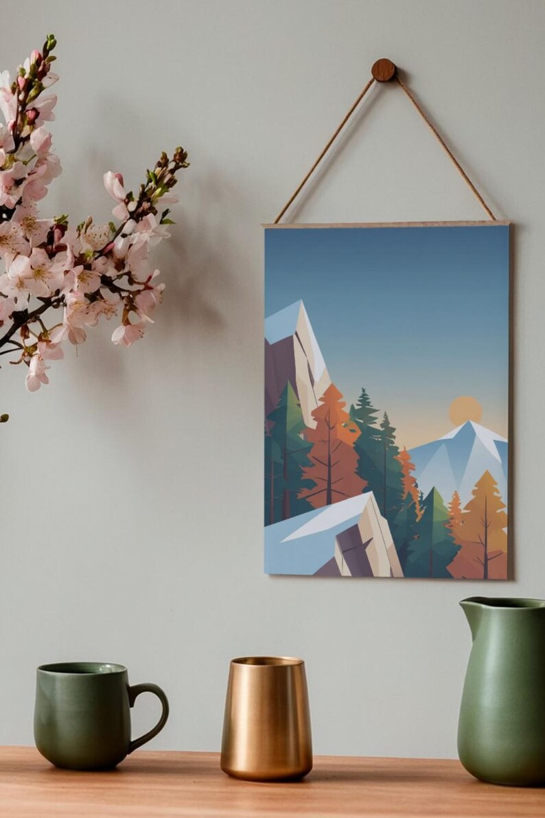 May include: A framed art print featuring a geometric mountain landscape with trees in shades of green, orange, and brown. The artwork is suspended by a wooden dowel and a natural cord. The background is a gradient of blue and the sun is setting.